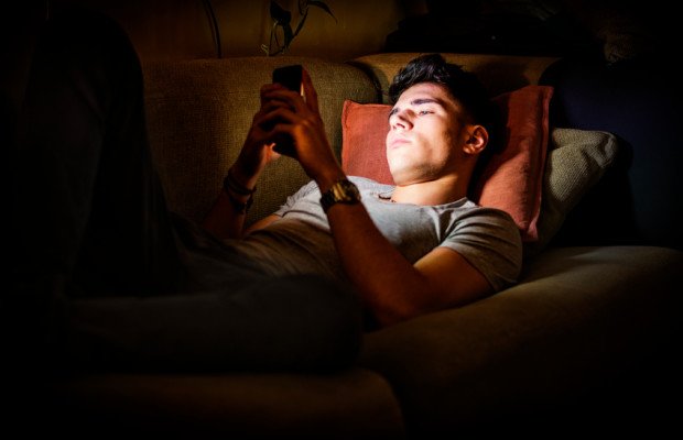 Attractive Young Man Lying on Sofa at Night and Illuminated by Light from Screen of Cell Phone at Home in Living Room