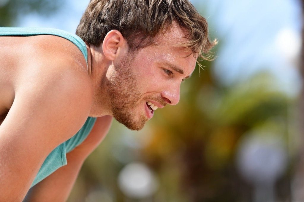 Tired exhausted man runner sweating after cardio workout. Running male adult taking a break and breaking a sweat after a run under the sun. Fitness athlete breathing heavily from heat exhaustion.