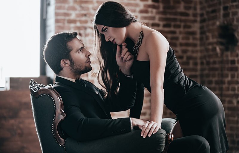 Beautiful young woman in cocktail dress leaning to her boyfriend sitting in chair while looking at each other in loft interior
