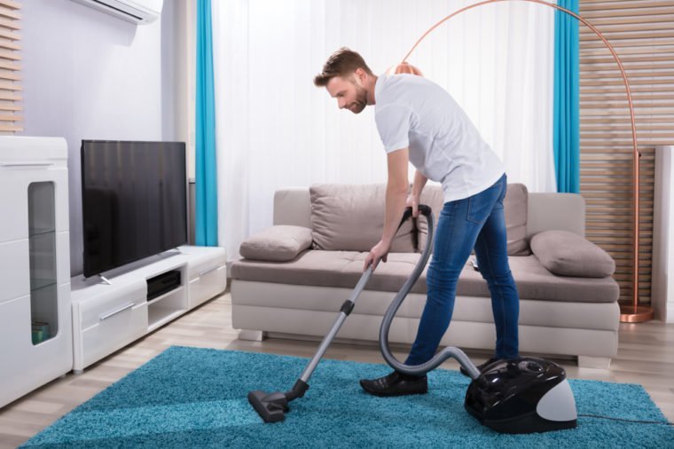 Young Man Cleaning Blue Carpet With Vacuum Cleaner At Home