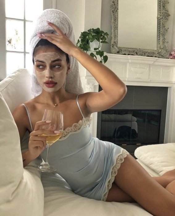 Chica bebiendo vino blanco, recostada en un sofá, usando mascarillas y con una toalla enredada en la cabeza