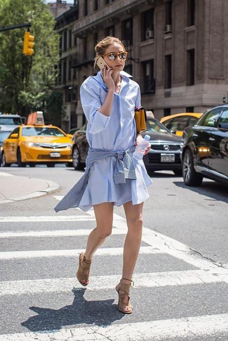 Chica caminando por la calle mientras habla por su celular, con vestido de camisa azul, camisa de cuadros amarrada a su cintura y gafas reflejantes
