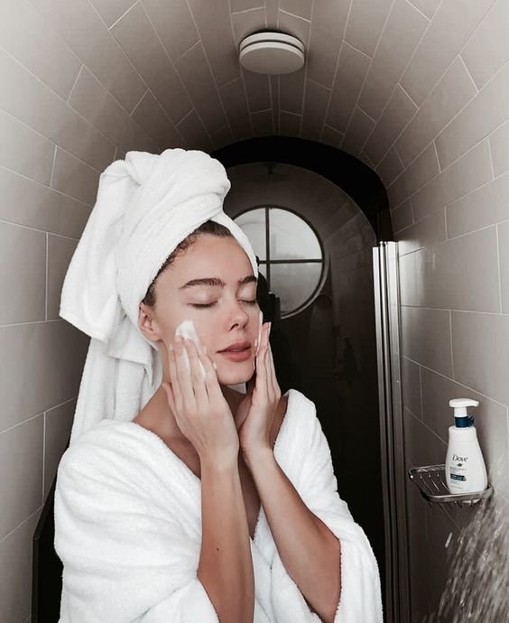 Chica en bata para baño color blanco con una toalla sobre su cabello, limpiando su rostro con jabón espumoso