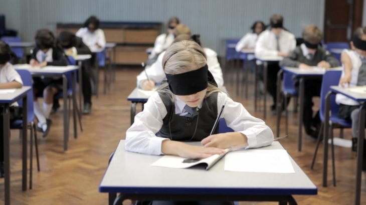niños de primaria sentados en pupitres con vendas en los ojos, haciendo un examen 