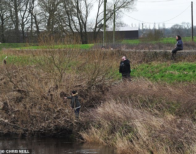 A member of the public today appears to point towards a spot in the undergrowth by the river Wyre while police watch on